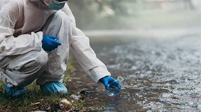 A scientist in a protective suit kneeling down to take a water sample from the river.