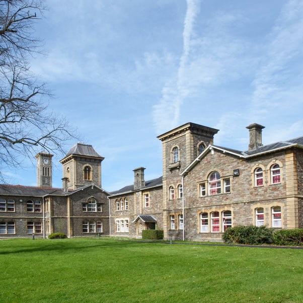 Exterior photograph of a large stone building seen through the gardens and magnolia tree at UWE Bristol Glenside Campus.
