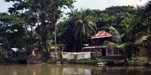 Image of a two storey house with lush greenery situated by a river bank in Bangladesh.