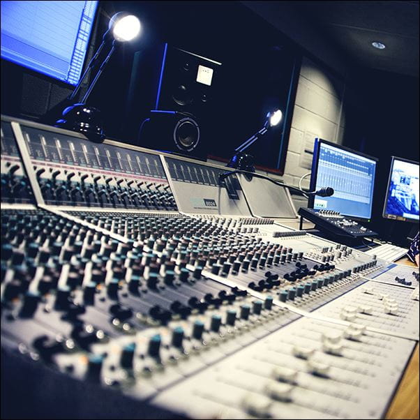 The interior of a sound recording studio with a large panel of control buttons with multiple computer screens.