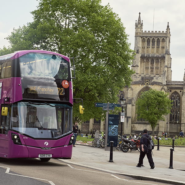 View of Bristol Cathedral with pedestrians and a First bus on the UWE Frenchay route in the foreground. There are trees and a bike park.