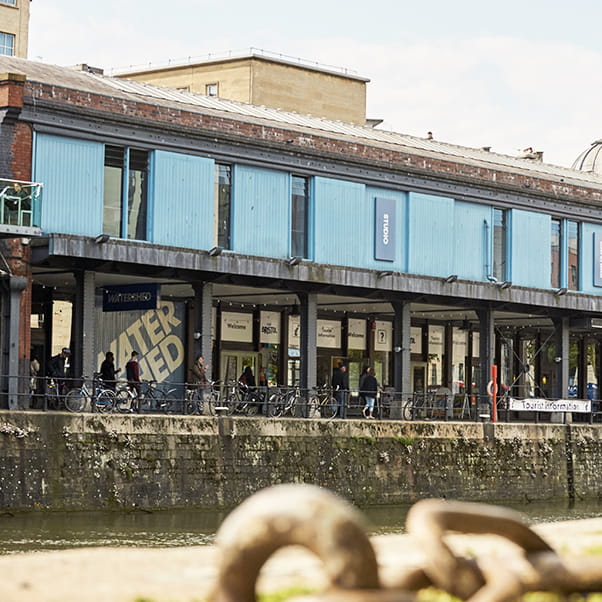 Image of The Watershed at the Bristol Harbourside in the daylight with a metal chain in the foreground and people walking past in the background.