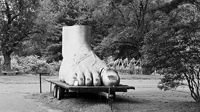 A black and white image of a right foot and ankle sculpture on a raised platform on a field surrounded by trees.