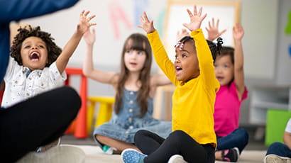 Image of a multi-ethnic group of preschool students sitting with their legs crossed on the floor in their classroom. The mixed-race female teacher is sitting on the floor facing the children. The happy kids are smiling and following the teacher's instructions. They have their arms raised in the air.