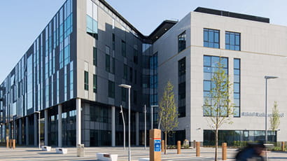 Photograph of the Bristol Business School building with paved terrace around it against a blue sky with a blurred cyclist riding by on the road.