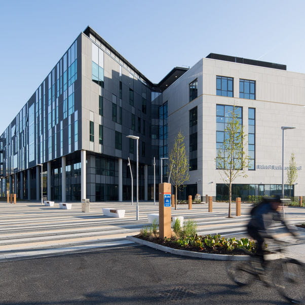 Photograph of the whole Bristol Business School building with paved terrace around it against a blue sky with a blurred cyclist riding by on the road.