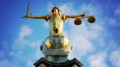 Upward angle shot of the Lady of Justice statue, holding a sword on her right hand and a pair of weighing scales on her left hand, at the top of the Old Bailey dome against a blue cloudy sky.