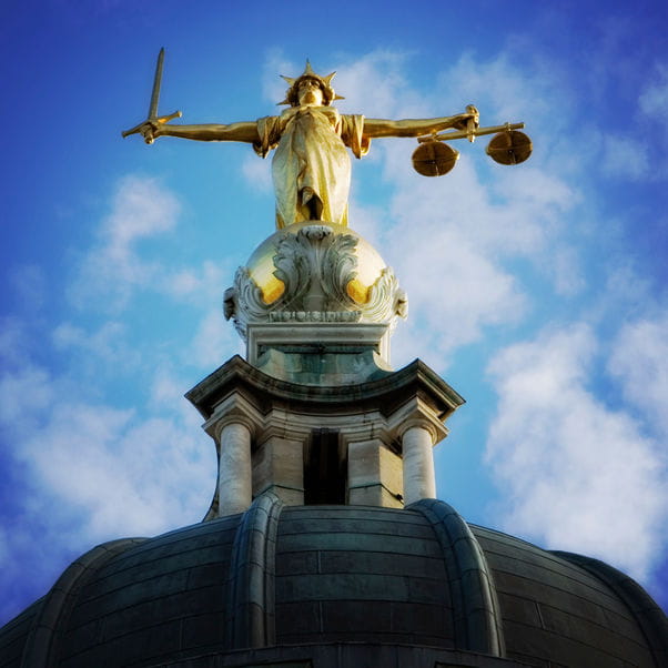 Upward angle shot of the Lady of Justice statue, holding a sword on her right hand and a pair of weighing scales on her left hand, at the top of the Old Bailey dome against a blue cloudy sky.