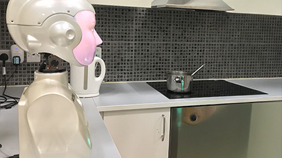 An assistive-living robot with a head and torso on a kitchen counter top in a kitchen. 