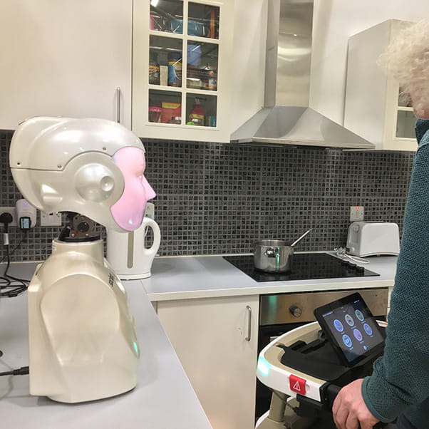An assistive-living robot with a head and torso on a kitchen counter top facing an elderly female with a walking frame in her kitchen. 