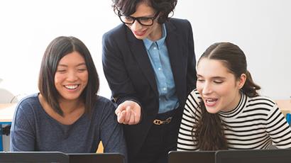 A group of three women, two women are seated with laptops whilst the third woman, who is the teacher, is showing referring to something on the computer screen.