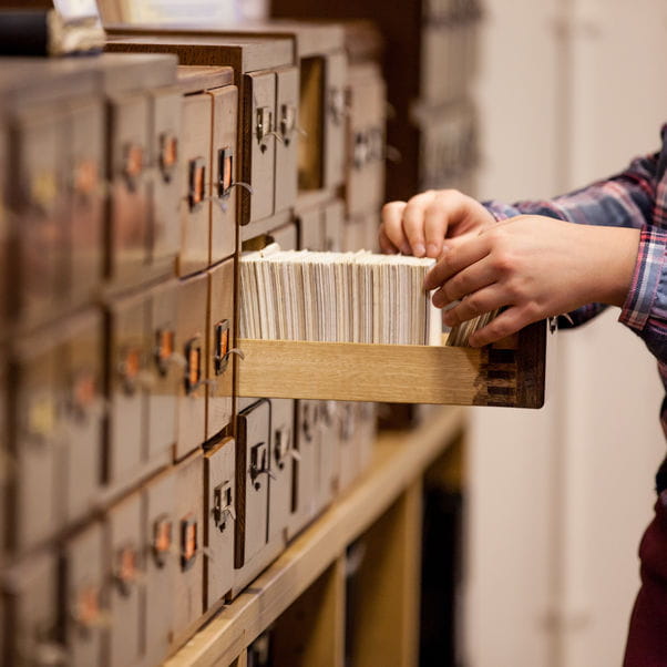 Image of filing card drawers with one open, and a pair of hands seeking a card.