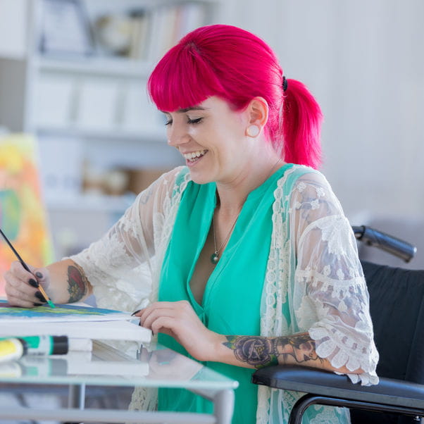 Young woman with pink hair working on a painting while sitting in a wheelchair.
