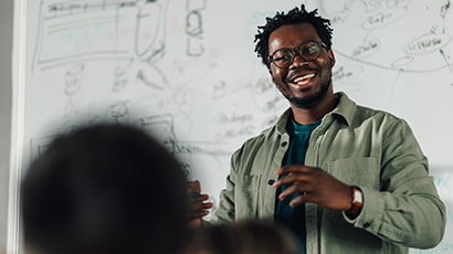 Lecturer in front of a white board, smiling as he looks toward a person in the classroom.