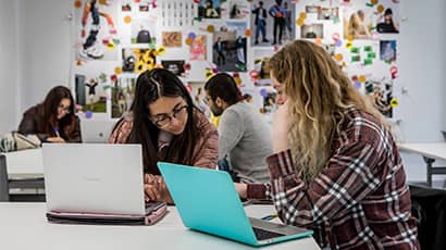 Two students at a desk discussing something on a laptop screen in a workspace in Bower Ashton.