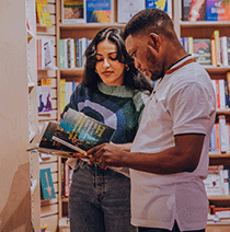 Two students standing together looking at a textbook in the library.