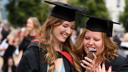 Two smiling students in graduation gowns looking at a phone