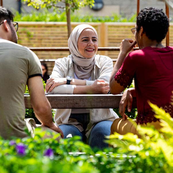 Couple of people chatting whilst sat around a table in a garden surrounded by shrubs. 
