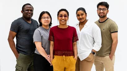 A group of smiling global majority students stand together. The person at the centre is wearing bright yellow linen trousers and a red top and the others are in neutrals.