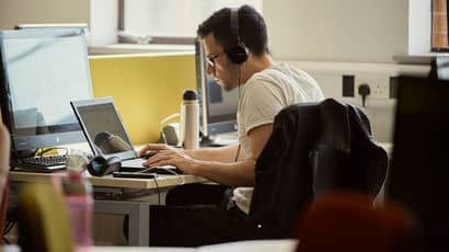 A person works on a laptop whilst wearing headphones.