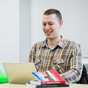 Man smiling whilst looking at a laptop on a desk with a few books.