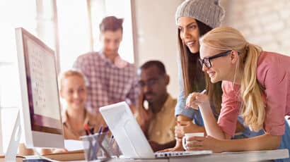 A group of students is gathered around a monitor and a laptop in a bright workspace.