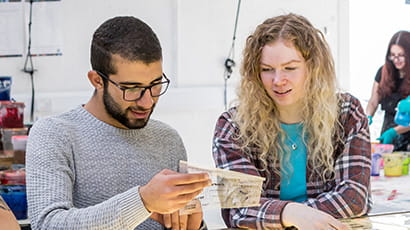 A group of three students sat at a table in a classroom setting, examining some art materials.