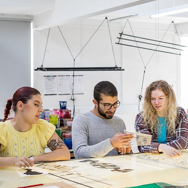 A group of three students sat at a table in a classroom setting, examining some art materials.