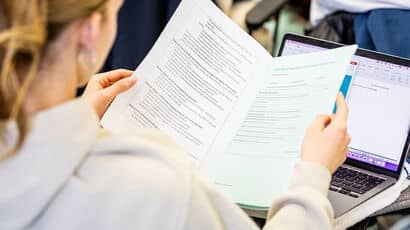 A young woman looking over some notes while working on a laptop.
