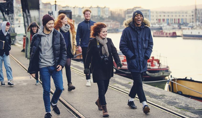 A group of students walk by the harbour in warm coats and hats. 
