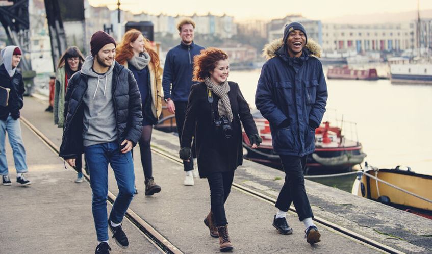 A group of students walk by the harbour in warm coats and hats.