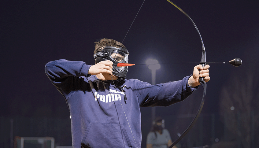 Man in sweatshirt and safety goggles pulling back on a bow to take aim.