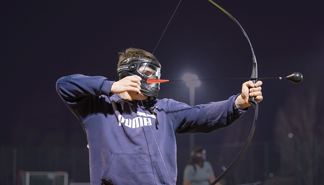 Man in sweatshirt and safety goggles pulling back on a bow to take aim.