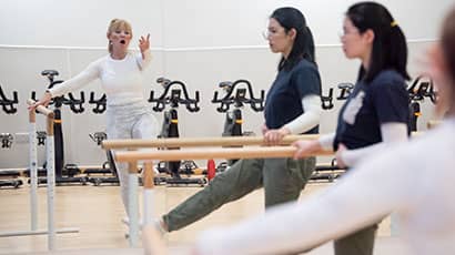 A student stands at the bar in front of a mirror with an instructor demonstrating a leg movement in the mirror reflection.