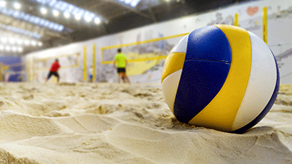 A volleyball nestled into the sand of volleyball pitch with players in the distance.