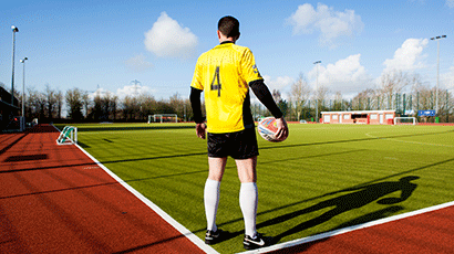 Man in bright yellow football shirt, holding a ball by his side at the corner of an astroturf football pitch.