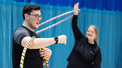 A student spins a hula hoop in the crook of his bent arm while a fellow student looks on smiling while she spins a hoop over her head.