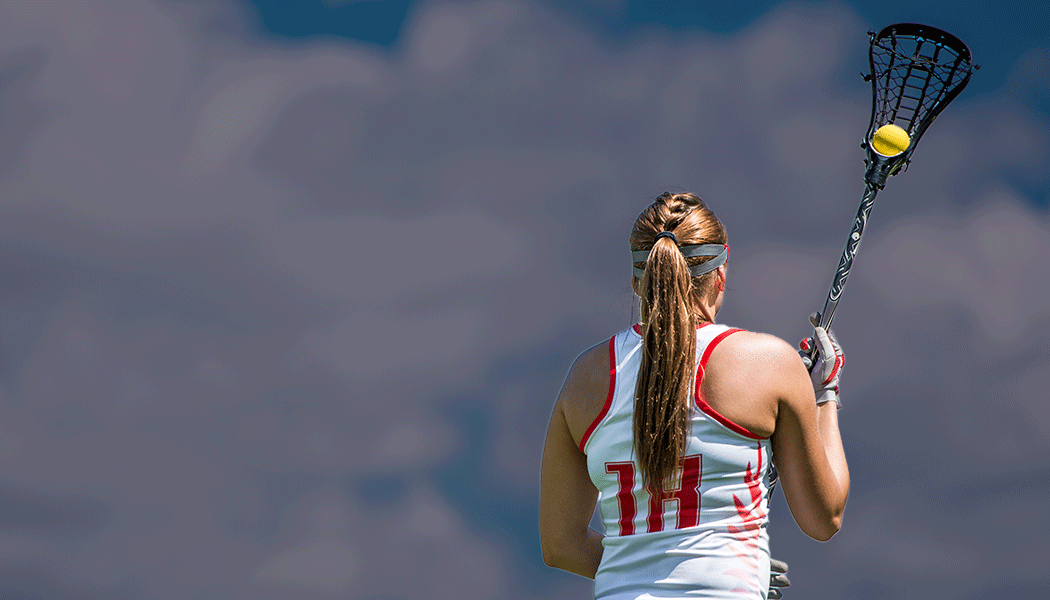 A lacrosse players stands with her back to camera, holding a lacrosse stick and ball high in front of her.