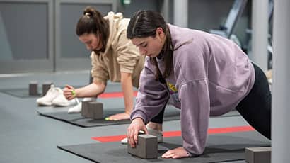 Two students on mats in yoga studio, one adjusting a block while in a low lunge.