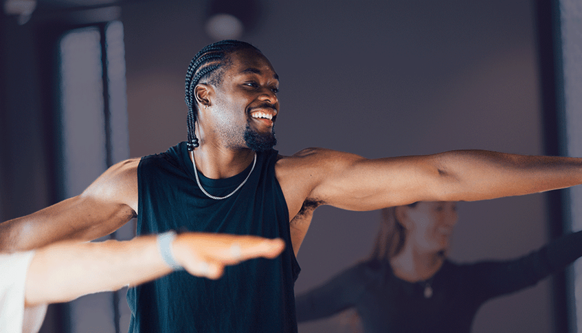 A man in the exercise studio, holding a warrior yoga pose and smiling.