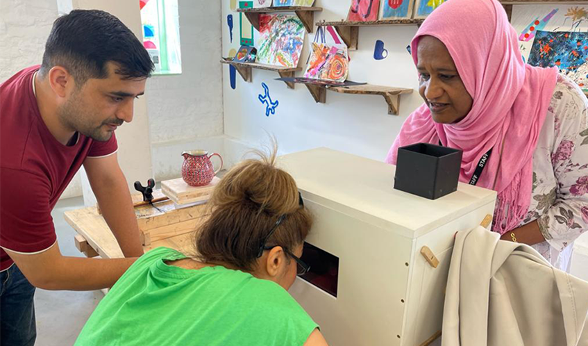 artists in a studio surrounding a white box on a table, which is part of a project. one of the artists is bending down and peering into the box through a small square cut into its side.