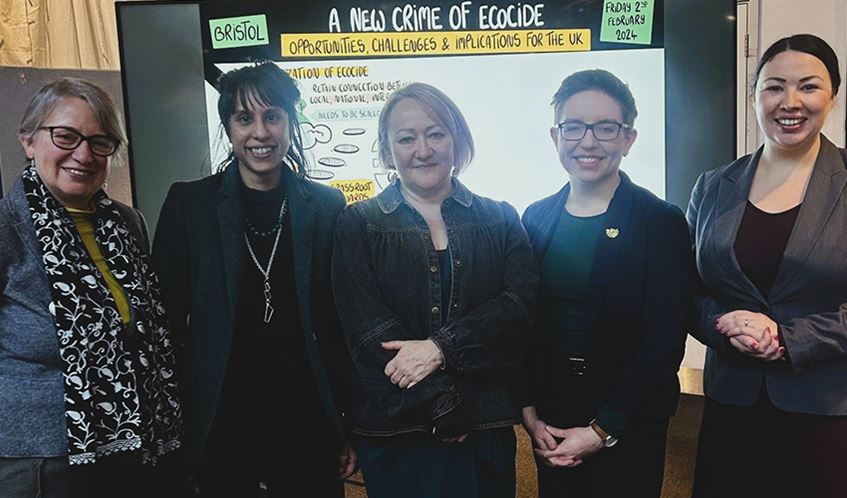 Five women smiling for a photo in front of a digital whiteboard that says 'A New Crime of Ecocide'