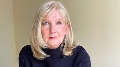 A woman with blonde hair and a black jumper looking at the camera against a beige background