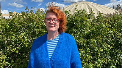 A woman stands in an outdoor setting in front of a green hedge looking directly at the camera and smiling